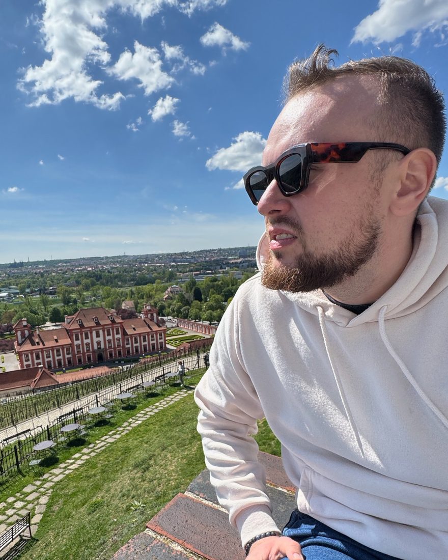 Aleš Prokop sitting on a wall above a château and its gardens, with the city on the horizon